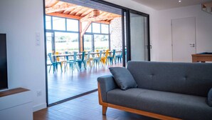 Una sala de estar moderna con un sofá gris, piso de madera y un amplio ventanal corredera que da a un área de comedor al aire libre con sillas coloridas.