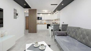 Una sala de estar moderna con un sofá gris, una mesa de centro con tazas, un televisor y una cocina al fondo.