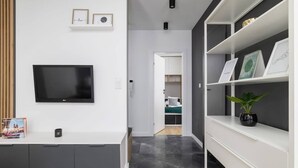 En moderne stue med et fladskærms-tv, et hvidt konsolbord med et magasin, og en reol med indrammede billeder.