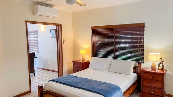 A bedroom with a bed, two bedside tables with lamps, a window with blinds, and an air conditioning unit.