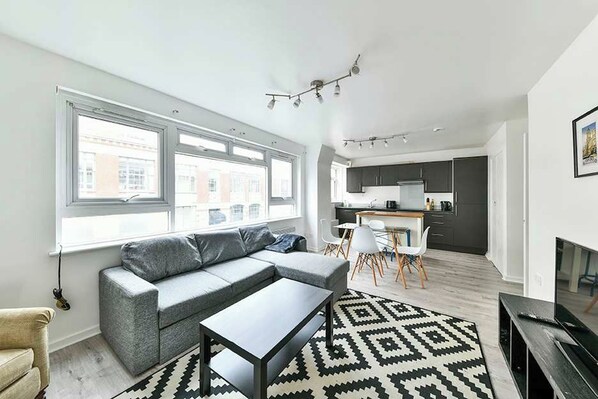 A modern living room with a grey sofa, a coffee table, and a patterned rug. There is a kitchen area with dark cabinets and a dining table with chairs. - The Elden - No.5 (Greater London)