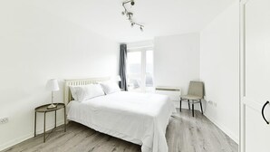 Une chambre à coucher au style minimaliste, avec un grand lit, une table de chevet avec une lampe, une chaise et une fenêtre avec des rideaux.