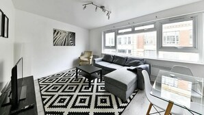 Un salon moderne avec un canapé, un pouf et une table en verre. On y trouve également un téléviseur, un fauteuil et un tapis à motifs.