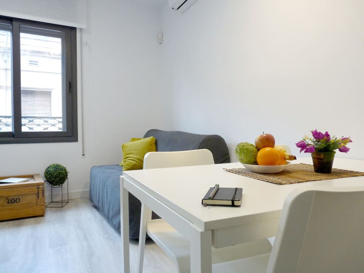 Una stanza piccola e moderna con un tavolo bianco, un divano grigio, una piccola pianta e una ciotola di frutta sul davanzale.