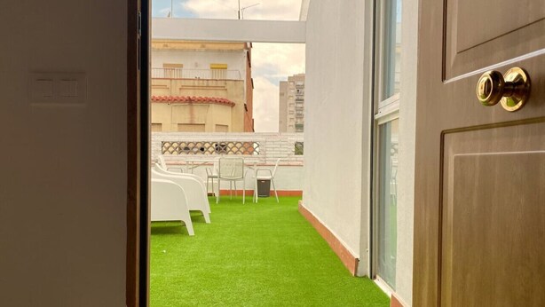 Vista de un balcón con césped artificial, sillas blancas y una mesa.