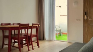 Un área de comedor con una mesa y sillas de madera, un divisor de vidrio corredizo que da a un espacio verde exterior, y una puerta de madera a la derecha.