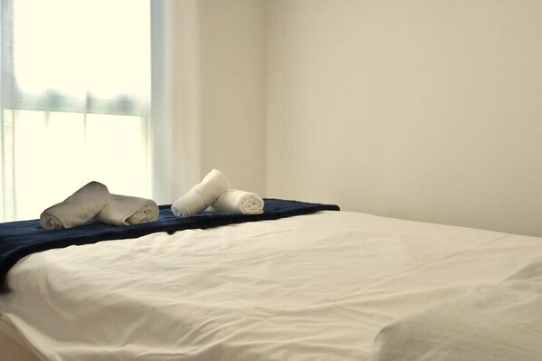 Una cama bien hecha con sábanas blancas y una manta azul oscuro, una ventana con cortinas traslúcidas y una pared blanca lisa.