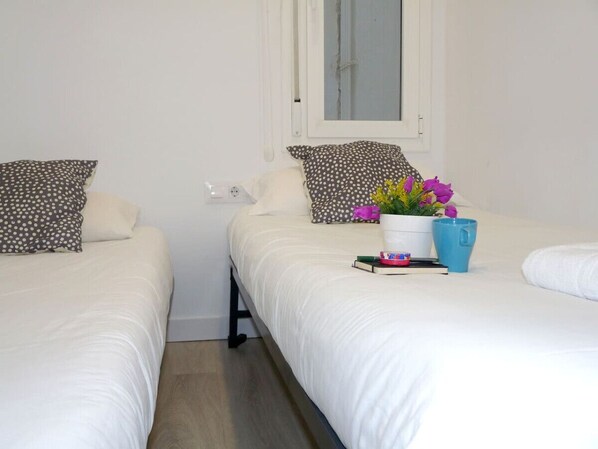 Habitación de hotel con dos camas, ropa de cama blanca, una ventana, una mesita de noche con una planta y un libro, y una taza azul.