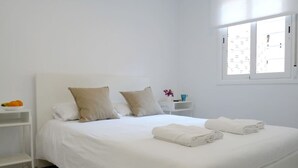 A neatly made bed with white linens and two pillows, a small bedside table with a cup and a flower vase, and a window with blinds.