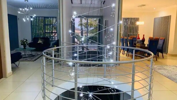 Un salon moderne doté d’une table ronde en verre entourée de rampes métalliques, d’un coin repas avec des chaises bleues et d’un escalier menant à un niveau supérieur.