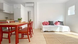 A modern, well-lit room with a wooden dining table and chairs, a bed with white bedding and pink pillows, and a large window providing natural light.