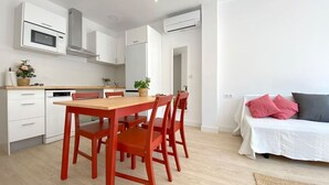 A modern kitchen with white cabinets, a wooden dining table, and red chairs. There is a white sofa with pink and gray pillows in the living area.