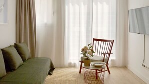 A room with a bed, a chair, a small table with a vase of flowers, and a TV on the wall.