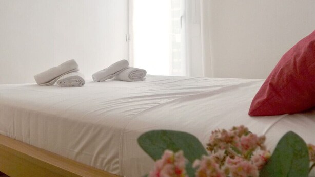 A neatly made bed with white linens and two rolled towels, a red pillow, and a window with sheer curtains.