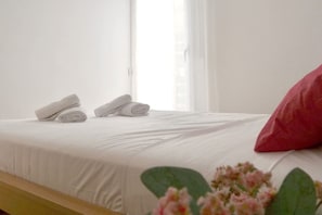 Una cama bien tendida con sábanas blancas, dos toallas enrolladas, un almohadón rojo y una ventana con cortinas traslúcidas.