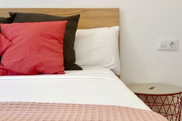 Una cama con cabecera de madera, dos almohadas, un colchón blanco, una mesita redonda con estructura roja y un enchufe en la pared.