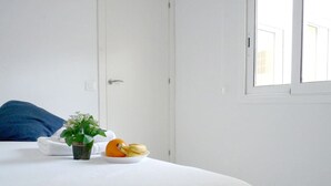 Ein ordentlich bezogenes Bett mit weißer Bettwäsche, ein kleiner Topfpflanze und ein Obstteller auf dem Nachttisch.