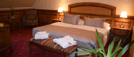 Habitación de hotel con una cama grande, dos mesitas de noche con lámparas, un banco de madera con toallas, una silla y una planta en maceta.