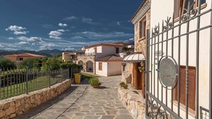 A stone-paved courtyard with a metal fence, stone walls, and a building with a tiled roof and wooden shutters. - Holiday apartment with swimming pool and air conditioning (Budoni)