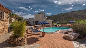 A pool area with lounge chairs and umbrellas, surrounded by stone walls and vegetation. - Holiday apartment with swimming pool and air conditioning (Budoni)