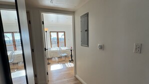 Un couloir doté d’un plancher en bois, de murs blancs et d’une bouche d’aération au plafond. Une porte donne accès à une pièce où se trouvent un lit et une chaise.