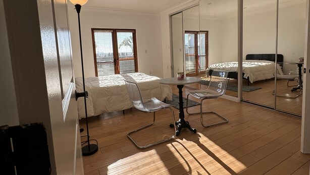 Une chambre moderne avec un grand lit, un bureau et une chaise. On y trouve une fenêtre avec des stores, une lampe et un miroir.