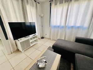 Una sala de estar con un televisor de pantalla plana sobre un mueble blanco, un sofá gris, una lámpara de pie y cortinas estampadas.