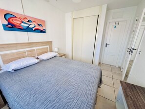 A neatly made bed with a grey blanket, a wooden headboard, and white pillows. There is a painting on the wall, a nightstand with a lamp, and a white door in the background. - A1047 - Nice apartment in a complete urbanisation and close to the beach (Villajoyosa)