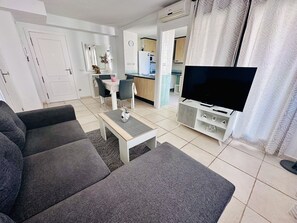 A living room with a grey sofa, a coffee table, a TV on a stand, and a kitchen in the background. - A1047 - Nice apartment in a complete urbanisation and close to the beach (Villajoyosa)