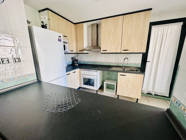 Cocina compacta con armarios de madera clara, encimera negra, refrigerador blanco, estufa y lavaplatos.
