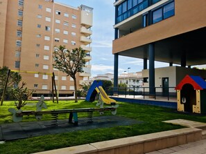 A playground with a slide and swings, surrounded by a grassy area and a building with multiple floors and balconies. - A1047 - Nice apartment in a complete urbanisation and close to the beach (Villajoyosa)