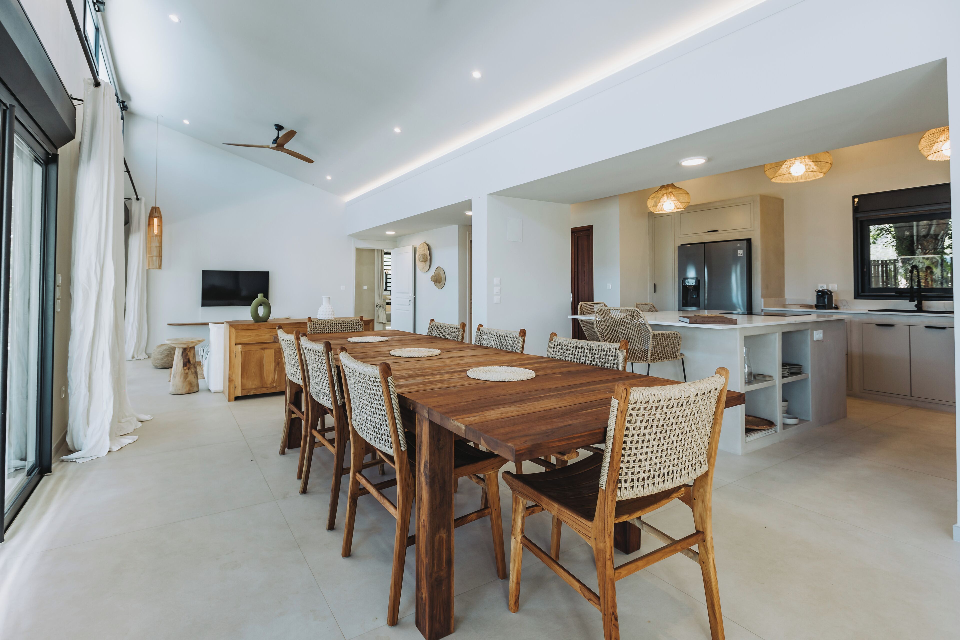 Un vaste espace dédié à la salle à manger, avec une longue table en bois et des chaises en osier, une cuisine équipée d’appareils électroménagers modernes et un ventilateur de plafond.