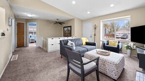 A spacious living room with a grey sofa, a dining table with a patterned table runner, and a large window with a view of the outside. - New! Recently Updated- Near Parks & Rec Center, Fenced Yard- 30 Day Rental (Littleton)