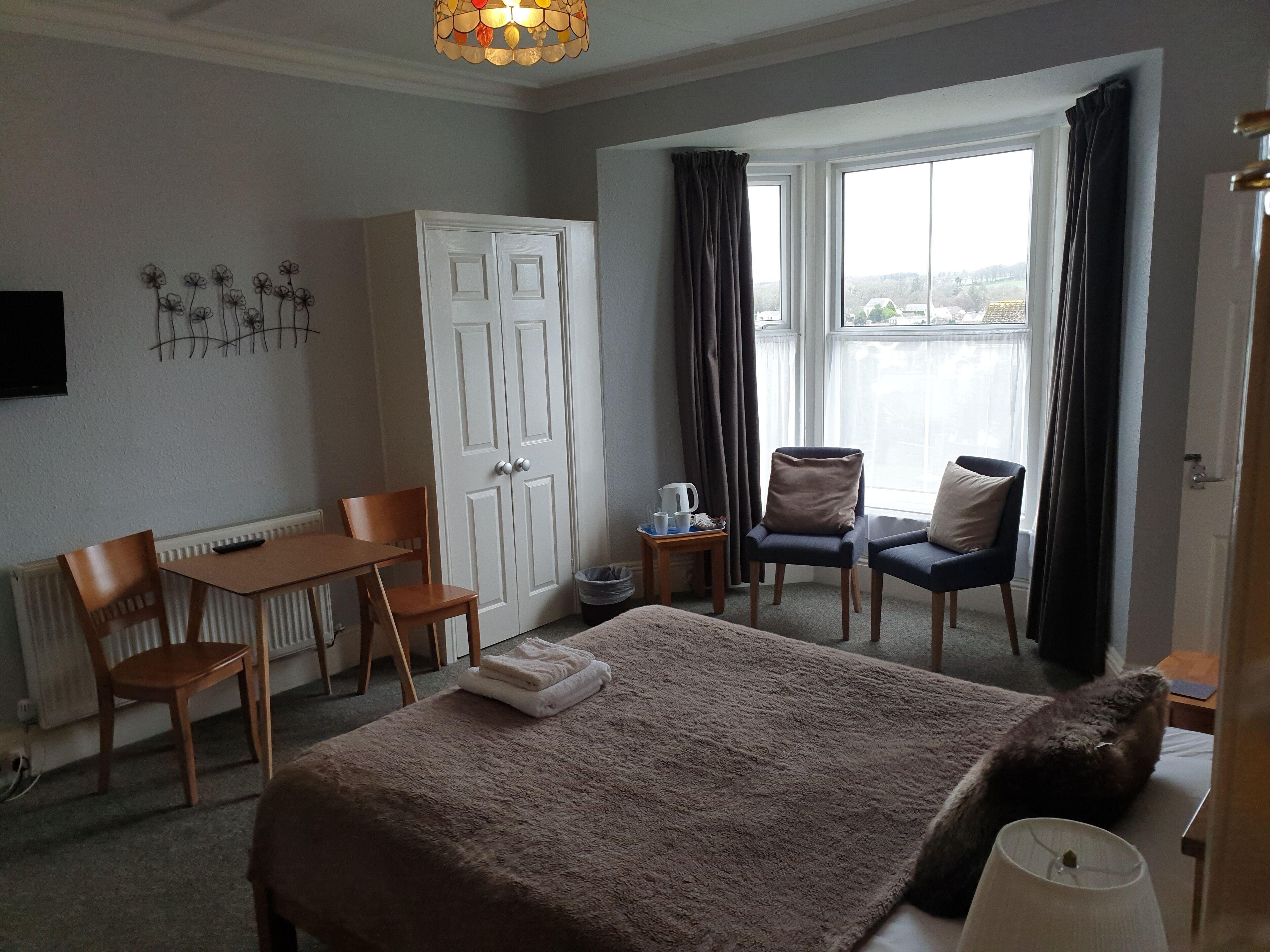 A hotel room with a bed, a desk, two chairs, a radiator, a window with curtains, and a wall-mounted TV.