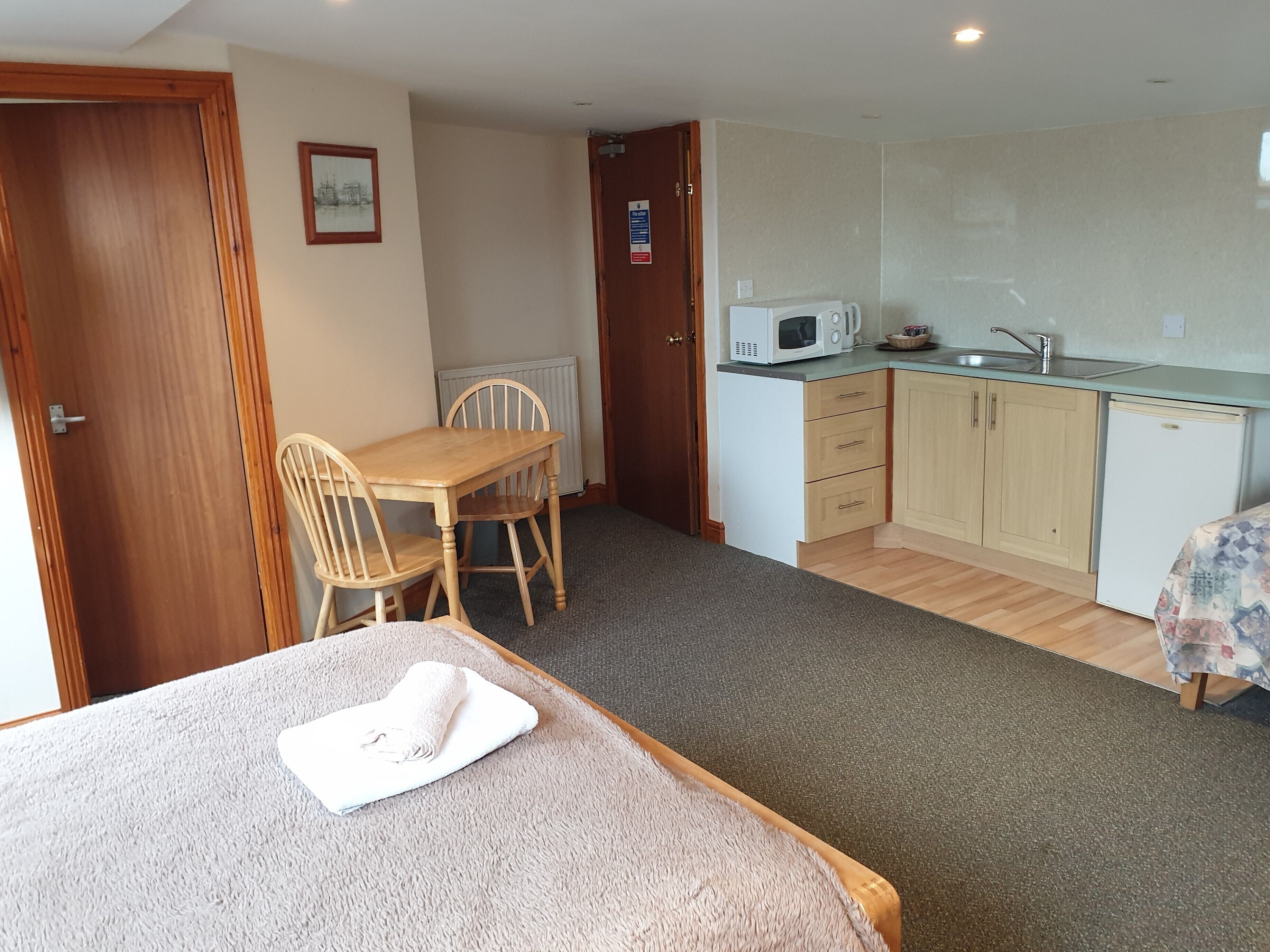 A compact kitchen with a microwave, sink, and wooden cabinets. There is a dining table with chairs and a small bed with a blanket.