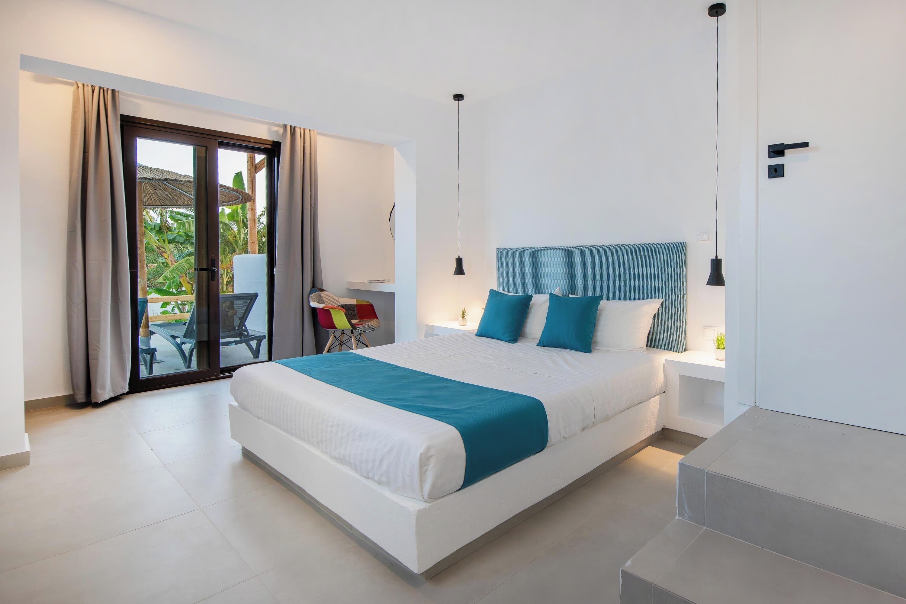 A modern hotel room with a large bed, a balcony with a chair, and a view of greenery.