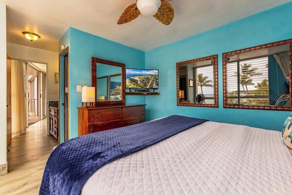 A bedroom with a large bed, a ceiling fan, a TV, and a view of palm trees through the window.