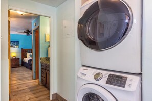 A laundry area with a Samsung washer and dryer, a wooden floor, and a bedroom visible through an open door. - Stunning Ocean View Gem with AC: Kamaole Nalu 504 - Your Island Getaway! (Kihei, Maui)