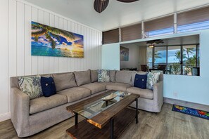 Ein Wohnzimmer mit einem Ecksofa, einem Glastisch und einem großen Gemälde einer tropischen Strandlandschaft.
