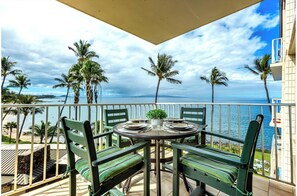 A balcony with a table set for two, overlooking a beach with palm trees and the ocean. - Stunning Ocean View Gem with AC: Kamaole Nalu 504 - Your Island Getaway! (Kihei, Maui)