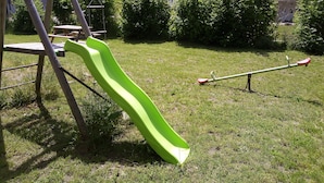 A playground with a slide and seesaw in a grassy yard with houses and trees in the background. - Nouveau Loft à Moins de 100m du lac Classé 2 (Gérardmer)