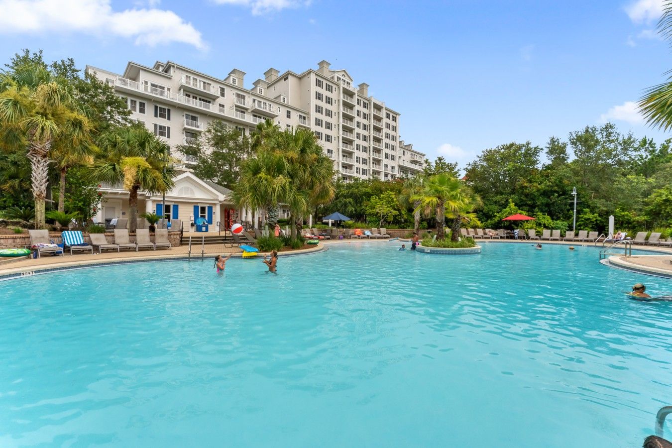 A swimming pool with lounge chairs and umbrellas, surrounded by palm trees and a multi-story building.