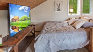 Ein Schlafzimmer mit Holzboden, einem Bett mit gemusterten Betttextilien, einem Fernseher auf einem Holzständer und einem Fenster mit Blick auf Grünflächen.