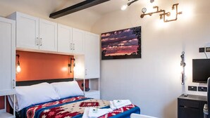 Hotellihuone, jossa on sänky, työpöytä, tuoli, televisio ja taulu, jossa on kuvattuna auringonlasku.