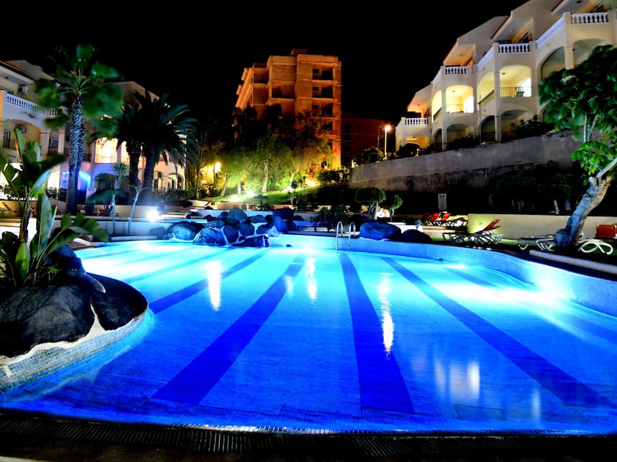 A swimming pool with blue lighting, surrounded by palm trees and a building with multiple balconies.