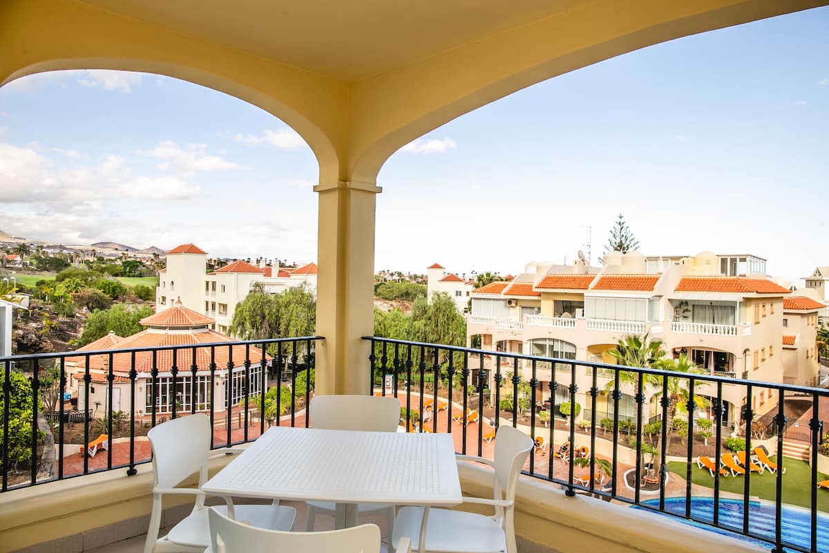 En balkon med et bord og stole, der ud til et poolområde og omkringliggende bygninger.