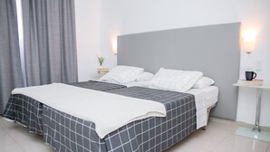 Un lit double avec une couvre-lit à carreaux gris et blancs, un oreiller blanc et une tête de lit grise. Il y a une table de chevet avec une tasse noire et une petite plante. La pièce est dotée d'un rideau gris et d'un mur blanc avec un interrupteur.