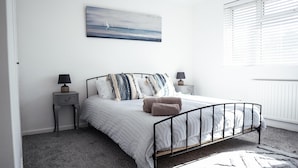 A bedroom with a bed, two bedside tables, a wall-mounted painting, and a window with blinds. - Inviting 4-bed House in Kettering (Kettering)