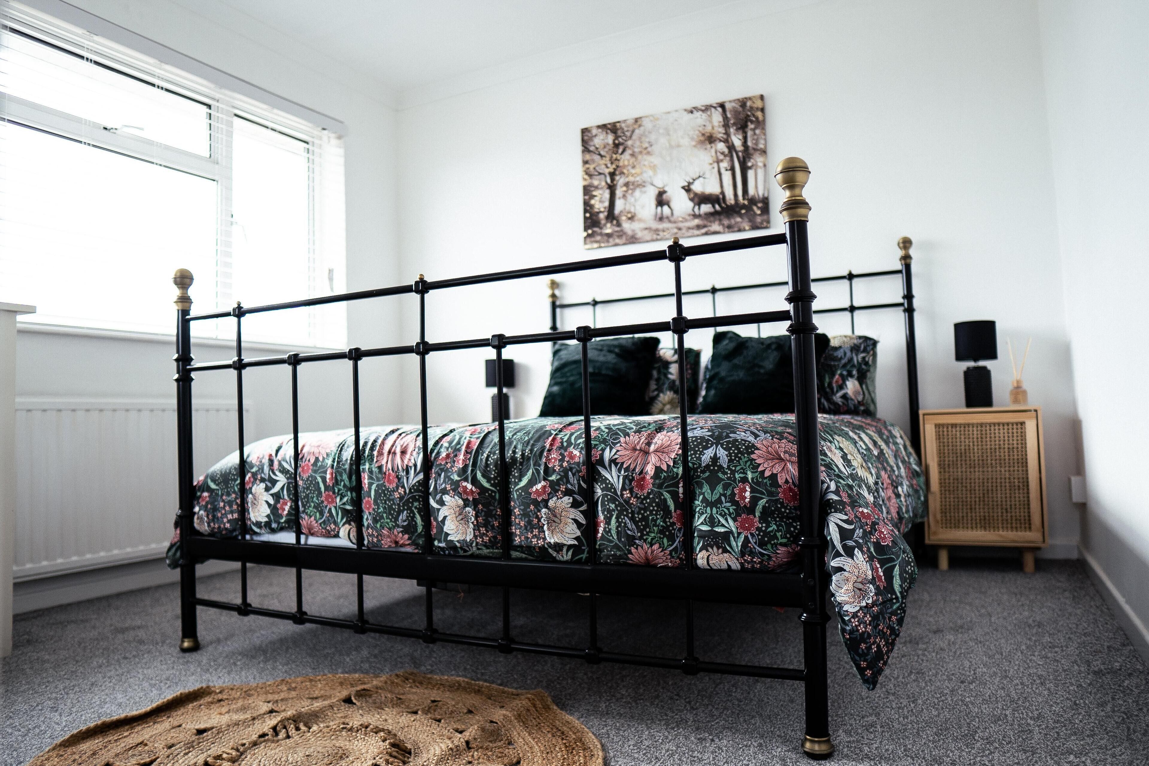 A bedroom with a black metal bed frame, floral bedding, a round woven rug, a painting of deer on the wall, and a small wooden cabinet with a lamp.