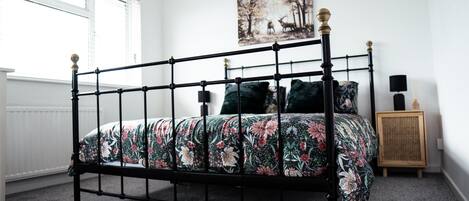 A bedroom with a black metal bed frame, floral bedding, a round woven rug, a painting of deer on the wall, and a small wooden cabinet with a lamp.